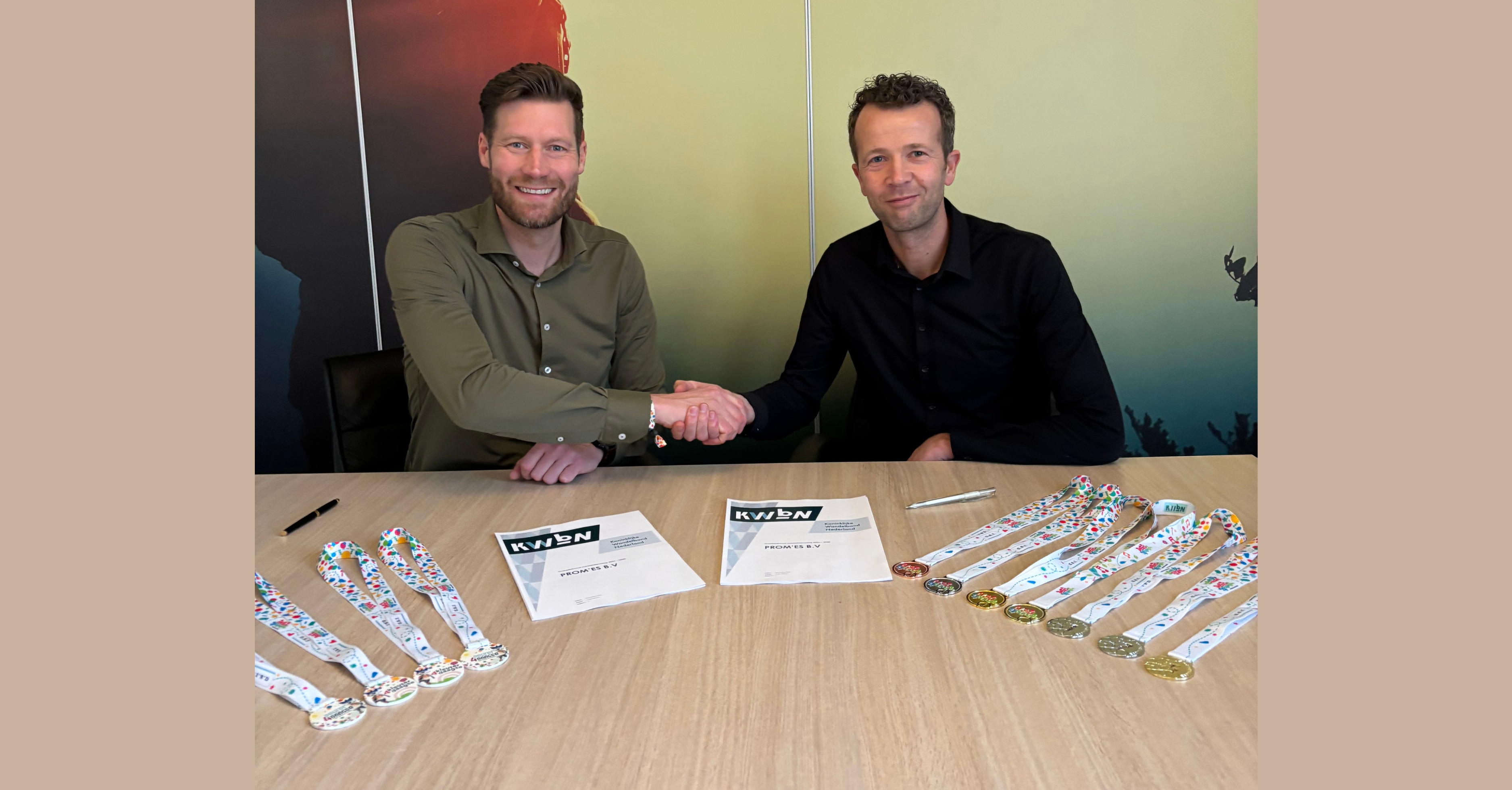 Twee mannen geven elkaar een hand. Op de voorgrond liggen twee contracten en Avond4daagse medailles